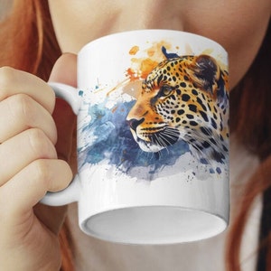 Könnte beinhalten: Weiße Keramiktasse mit einem Aquarelldesign eines Leopardenkopfes. Der Leopard ist braun und schwarz mit orangefarbenen Flecken. Der Hintergrund ist ein blauer und orangefarbener Aquarell-Wasch.