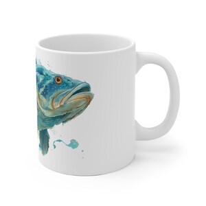 May include: White ceramic mug featuring a watercolor illustration of a blue fish with orange eyes. The fish has detailed scales and is set against a white background. The mug has a standard handle.