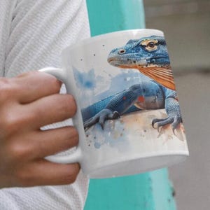 Może przedstawiać: Biały kubek ceramiczny z wzorem niebieskiego i pomarańczowego jaszczurki. Jaszczurka jest namalowana w stylu akwarelowym z rozbryzgami koloru.