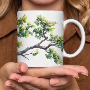 Może przedstawiać: Biały kubek ceramiczny z akwarelowym wzorem zielonej, liściastej gałęzi na białym tle.
