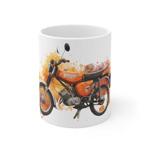 May include: White ceramic mug featuring a watercolor illustration of an orange Simson motorcycle. The design includes the word "Simson" and is set against a backdrop of orange and yellow paint splatters.