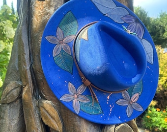 May include: A vibrant blue fedora hat with hand-painted floral and leaf designs in shades of gray, pink, and green. The hat features a brown leather band and is displayed outdoors.