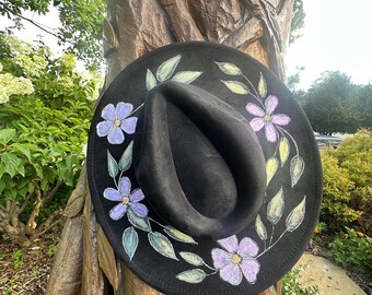 May include: A black fedora-style hat with a wide brim. The brim is decorated with hand-painted purple flowers and green leaves. The hat is displayed outdoors, resting on a tree trunk.