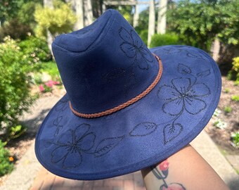 May include: A navy blue fedora-style hat with a wide brim. The hat features black floral embroidery and a brown braided band around the crown. The hat is made of a soft material and is suitable for sun protection.