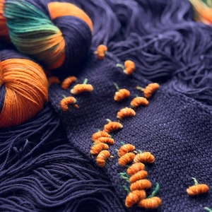 Könnte beinhalten: Ein dunkelviolettes Strickstück mit kleinen, gestickten orangefarbenen Kürbismotiven mit grünen Stielen. Der Stoff ist von Garnsträngen in passenden dunkelvioletten sowie melierten orangefarbenen, grünen und schwarzen Farbtönen umgeben.