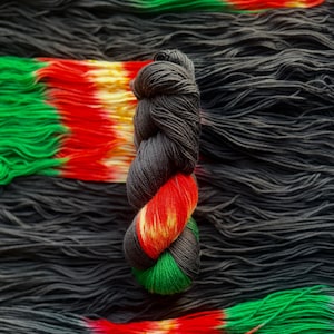 May include: A skein of hand-dyed yarn in shades of black, red, green, and yellow. The yarn is twisted into a compact bundle, with the colors transitioning along its length. The background features loose strands of black yarn and a colorful gradient.