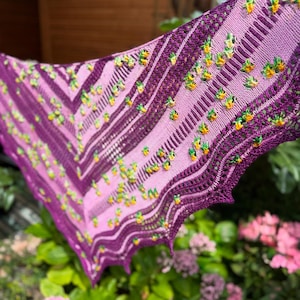 May include: A handmade, purple and pink shawl with a floral design. The shawl is knitted with a variety of stitches, creating a textured look. The edges are finished with a delicate lace pattern. The shawl is decorated with small, colorful flowers.