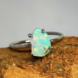 Può includere: Un anello d'argento con una vibrante pietra di opale dalla forma irregolare. L'opale mostra una miscela di colori iridescenti, tra cui blu, verdi e gialli. L'anello è posto su una superficie di legno.