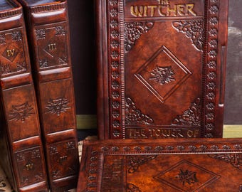May include: A collection of four brown, leather-bound books with intricate embossed patterns and metallic corner accents. One book features the title The Witcher and a wolf emblem. Two books are standing upright, while two are lying flat.
