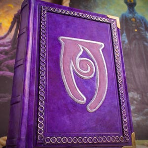 May include: A purple book with a textured cover featuring a central silver and light purple emblem. The cover is bordered by a silver chain-link pattern and includes gold-toned metal corner protectors. The spine shows raised horizontal bands.