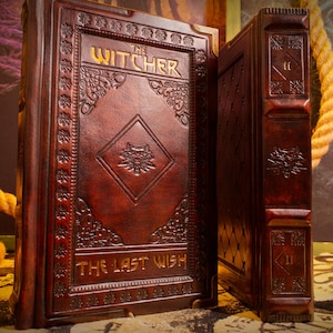 May include: Two leather-bound books with embossed designs. The book on the left is open, displaying the title "The Witcher" and "The Last Wish" in gold lettering. The cover features a wolf emblem. The books are a deep reddish-brown colour.
