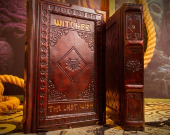 May include: Two leather-bound books with embossed designs. The book on the left is open, displaying the title "The Witcher" and "The Last Wish" in gold lettering. The cover features a wolf emblem. The books are a deep reddish-brown color.