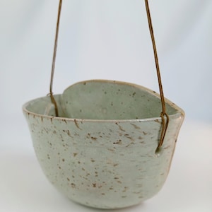 Könnte beinhalten: Ein hängender Keramik-Pflanztopf in Hellgrün mit braunen Sprenkeln. Der schalenförmige Topf hat einen geschwungenen Rand und wird von zwei braunen Kordeln gehalten. Minimalistisches Design, geeignet für kleine Pflanzen.