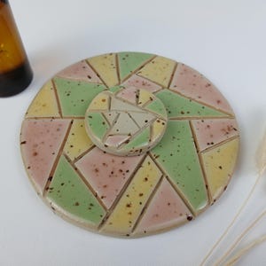 Könnte beinhalten: Ein Keramikuntersetzer mit geometrischem Design in Pastellfarben Rosa, Grün und Gelb. Ein kleinerer, passender Untersetzer liegt obenauf. Im Hintergrund befindet sich eine braune Glasflasche.