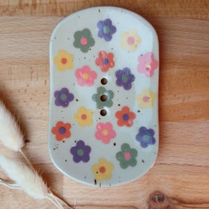 Könnte beinhalten: Eine Keramik-Seifenschale mit hellgrünem Hintergrund, verziert mit bunten Blumen in Rosa-, Gelb-, Orange-, Lila- und Grüntönen. Die Schale hat drei Abflusslöcher und eine rechteckige Form mit abgerundeten Kanten.