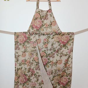 Könnte beinhalten: Schürze mit einem Blumenmuster aus rosa und pfirsichfarbenen Rosen auf beigem Hintergrund. Die Schürze hat eine Rüschenborte und hängt an einem Holzbügel. Die Bindebänder der Schürze sind einfarbig beige.