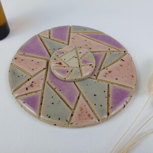 Könnte beinhalten: Ein Keramik-Untersetzer-Set mit geometrischem Design. Der große Untersetzer ist rund, mit einem Mosaikmuster aus grauen, rosa und lila Dreiecken. Ein kleinerer, passender Untersetzer liegt obenauf. Eine braune Flasche ist im Hintergrund.