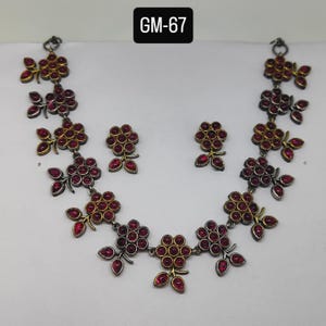 Puede incluir: Un conjunto de collar y pendientes con un diseño floral. El collar y los pendientes tienen una base de metal plateado con detalles dorados. Las flores están compuestas por pequeñas piedras preciosas rojas redondas. El texto "GM-67" está en la parte superior central.