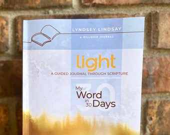 May include: A softcover journal titled "light" with the text "My Word in 30 Days" and "A Guided Journal Through Scripture." The cover features a watercolor design in shades of yellow and gold, with a blue and white grid pattern at the top. The journal is held against a brick wall.