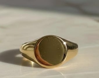 May include: A gold signet ring with a polished, circular face. The ring has a smooth, rounded band and a bright, reflective finish. The ring is set against a light-colored background.