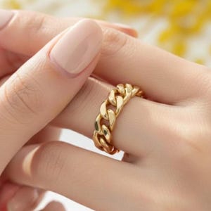 Può includere: Un anello a catena dorata. L'anello presenta un design a catena spessa e ad incastro. L'anello è mostrato su un dito, con una manicure dai toni neutri. Il design è elegante.
