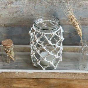 May include: A glass jar with a white macrame cover, a white candle inside, and a wooden tray with other glass jars filled with dried flowers.
