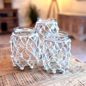 May include: Three clear glass jars with a white macrame netting design. The jars are arranged on a wooden surface, with a blurred background featuring furniture and a lamp. The jars are empty and ready to be filled.