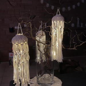 May include: Three white macrame hanging light fixtures with warm white LED lights inside. The lights are hanging from a branch and are displayed on a black surface.