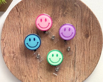 Puede incluir: Cuatro carretes de insignia con caras sonrientes de colores en rosa, azul, morado y verde. Cada carrete tiene un diseño de cara sonriente y un cordón retráctil. Los carretes están dispuestos sobre una superficie redonda de madera.