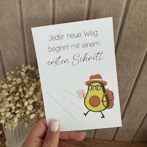 Könnte beinhalten: Weiße Karte mit dem deutschen Text "Jeder neue Weg beginnt mit einem ersten Schritt" und einem Cartoon-Avocado-Charakter mit Hut, Brille und Rucksack, der eine Karte hält. Die Avocado geht auf einer Straße.