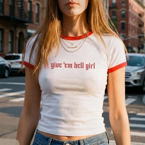 May include: White ringer t-shirt with red trim and the phrase "give 'em hell girl" in red lettering. The shirt is paired with blue jeans. The Smooth Rebellion Company original design.