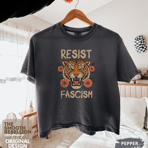 RESIST FASCISM Tiger Boxy T-Shirt for women. Political Protest Graphic Tee. Boxy tee that features a vintage-style painted tiger surrounded by vibrant poppies. Perfect for protests, marches, or just everyday defiance. Political merch for making a statement. Anti Trump top made for the loud and the politically engaged. Whether you're fighting fascism, defending democracy, or just making a statement. Short sleeves, crew neck. Boxy cropped tee. Very soft fabric. Printed in the US.