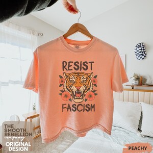 RESIST FASCISM Tiger Boxy T-Shirt for women. Political Protest Graphic Tee. Boxy tee that features a vintage-style painted tiger surrounded by vibrant poppies. Perfect for protests, marches, or just everyday defiance. Political merch for making a statement. Anti Trump top made for the loud and the politically engaged. Whether you're fighting fascism, defending democracy, or just making a statement. Short sleeves, crew neck. Boxy cropped tee. Very soft fabric. Printed in the US.