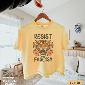 RESIST FASCISM Tiger Boxy T-Shirt for women. Political Protest Graphic Tee. Boxy tee that features a vintage-style painted tiger surrounded by vibrant poppies. Perfect for protests, marches, or just everyday defiance. Political merch for making a statement. Anti Trump top made for the loud and the politically engaged. Whether you're fighting fascism, defending democracy, or just making a statement. Short sleeves, crew neck. Boxy cropped tee. Very soft fabric. Printed in the US.