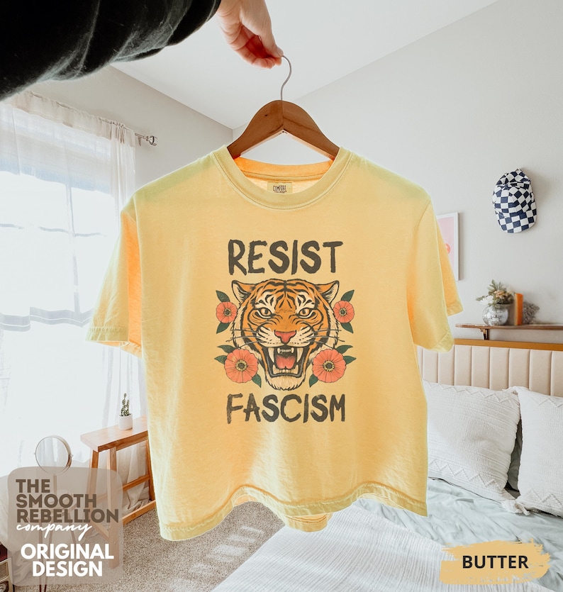 RESIST FASCISM Tiger Boxy T-Shirt for women. Political Protest Graphic Tee. Boxy tee that features a vintage-style painted tiger surrounded by vibrant poppies. Perfect for protests, marches, or just everyday defiance. Political merch for making a statement. Anti Trump top made for the loud and the politically engaged. Whether you're fighting fascism, defending democracy, or just making a statement. Short sleeves, crew neck. Boxy cropped tee. Very soft fabric. Printed in the US.