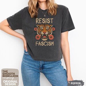 RESIST FASCISM Tiger Boxy T-Shirt for women. Political Protest Graphic Tee. Boxy tee that features a vintage-style painted tiger surrounded by vibrant poppies. Perfect for protests, marches, or just everyday defiance. Political merch for making a statement. Anti Trump top made for the loud and the politically engaged. Whether you're fighting fascism, defending democracy, or just making a statement. Short sleeves, crew neck. Boxy cropped tee. Very soft fabric. Printed in the US.