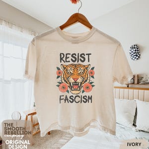 RESIST FASCISM Tiger Boxy T-Shirt for women. Political Protest Graphic Tee. Boxy tee that features a vintage-style painted tiger surrounded by vibrant poppies. Perfect for protests, marches, or just everyday defiance. Political merch for making a statement. Anti Trump top made for the loud and the politically engaged. Whether you're fighting fascism, defending democracy, or just making a statement. Short sleeves, crew neck. Boxy cropped tee. Very soft fabric. Printed in the US.