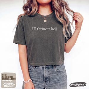 May include: A charcoal gray short-sleeve t-shirt with a vintage-washed finish. The shirt features the text "I'll thrive in hell" printed in a white, gothic-style font across the chest. The garment has a crew neckline and a relaxed, boxy fit.