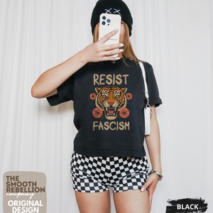 RESIST FASCISM Tiger Boxy T-Shirt for women. Political Protest Graphic Tee. Boxy tee that features a vintage-style painted tiger surrounded by vibrant poppies. Perfect for protests, marches, or just everyday defiance. Political merch for making a statement. Anti Trump top made for the loud and the politically engaged. Whether you're fighting fascism, defending democracy, or just making a statement. Short sleeves, crew neck. Boxy cropped tee. Very soft fabric. Printed in the US.