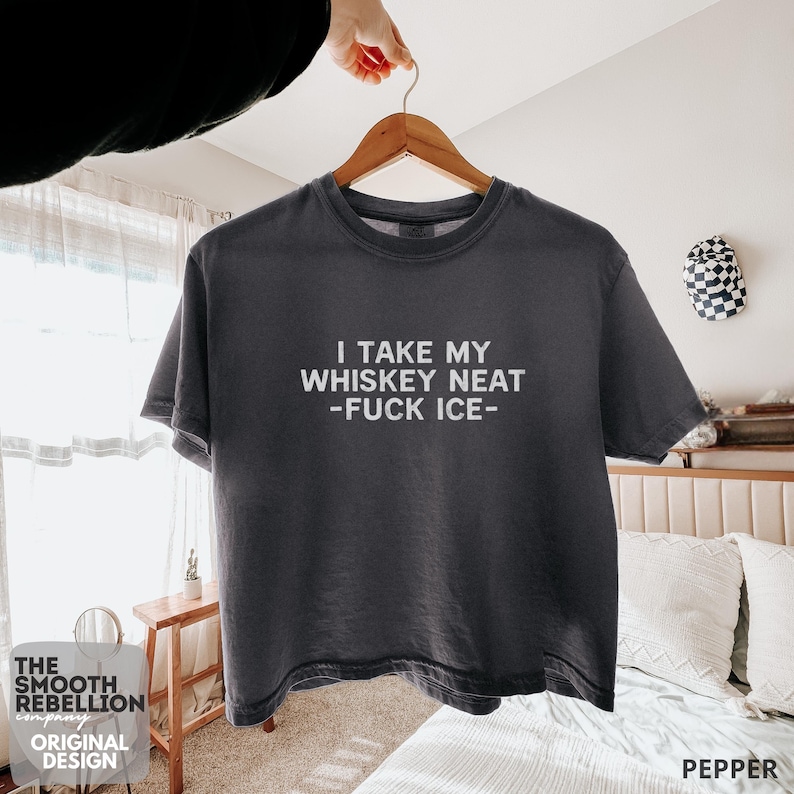 Pepper Comfort Colors boxy tee. The shirt has bold black text reading “I TAKE MY WHISKEY NEAT – FUCK ICE –.” Slightly Cropped. Boxy, relaxed fit. 100% Cotton. Heavy Fabric. Protest & Anti Ice Apparel.