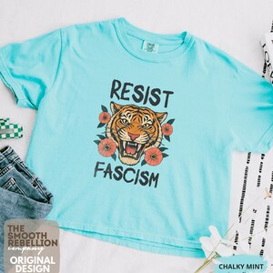 RESIST FASCISM Tiger Boxy T-Shirt for women. Political Protest Graphic Tee. Boxy tee that features a vintage-style painted tiger surrounded by vibrant poppies. Perfect for protests, marches, or just everyday defiance. Political merch for making a statement. Anti Trump top made for the loud and the politically engaged. Whether you're fighting fascism, defending democracy, or just making a statement. Short sleeves, crew neck. Boxy cropped tee. Very soft fabric. Printed in the US.