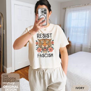RESIST FASCISM Tiger Boxy T-Shirt for women. Political Protest Graphic Tee. Boxy tee that features a vintage-style painted tiger surrounded by vibrant poppies. Perfect for protests, marches, or just everyday defiance. Political merch for making a statement. Anti Trump top made for the loud and the politically engaged. Whether you're fighting fascism, defending democracy, or just making a statement. Short sleeves, crew neck. Boxy cropped tee. Very soft fabric. Printed in the US.