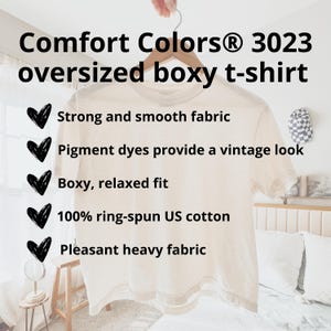 May include: A cream-colored Comfort Colors 3023 oversized boxy t-shirt hanging on a wooden hanger. The shirt features the text: "Strong and smooth fabric", "Pigment dyes provide a vintage look", "Boxy, relaxed fit", "100% ring-spun US cotton", and "Pleasant heavy fabric".