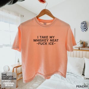 Peachy Comfort Colors boxy tee. The shirt has bold black text reading “I TAKE MY WHISKEY NEAT – FUCK ICE –.” Slightly Cropped. Boxy, relaxed fit. 100% Cotton. Heavy Fabric. Protest & Anti Ice Apparel.