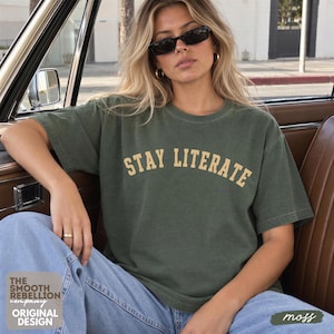 May include: A short-sleeved olive green crewneck t-shirt featuring the text STAY LITERATE printed in a curved, light yellow collegiate-style font across the chest. The shirt is made of a solid-colored fabric with a standard fit and hem.