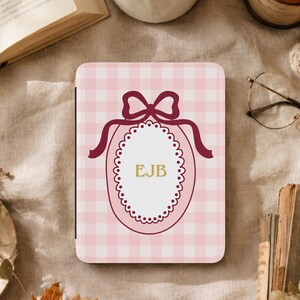 May include: A rectangular e-reader case featuring a light pink and white gingham pattern. The center displays a white oval with a scalloped border, topped with a dark red bow graphic. The letters EJB are printed in gold in the center.