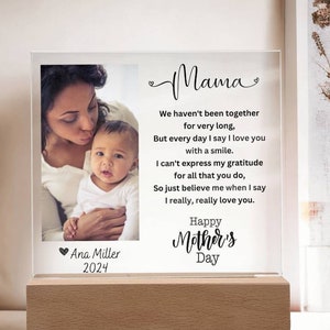 May include: A clear acrylic photo frame with a wood base. The frame holds a photo of a mother and child. The frame has a message that reads "Mama We haven't been together for very long, But every day I say I love you with a smile. I can't express my gratitude for all that you do, So just believe me when I say I really, really love you. Happy Mother's Day" and is signed "Ana Miller 2024".