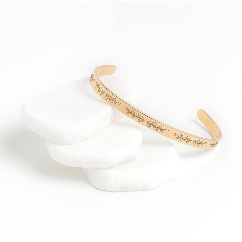 May include: A gold-toned bangle bracelet with a delicate leaf design engraved around the band.
