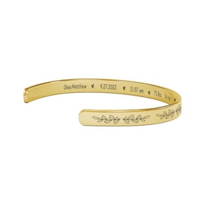 May include: Gold bangle bracelet with an engraved inscription: "Olivia Matthew &hearts; 9.27.2022 &hearts; 11:37 am &hearts; 7 lbs 14 oz" and a leafy design.