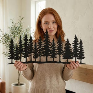 Op de afbeelding: Zwart metalen wandkunst met een bos van dennenbomen. Het silhouetontwerp toont meerdere bomen van verschillende hoogtes, waardoor een gelaagd effect ontstaat. Het kunstwerk wordt door een persoon vastgehouden.
